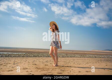 Isolierte Frau im Blumenkleid, die in den Sommerferien am britischen Strand entlang läuft und ihren großen Strohsonnenhut hält. Stockfoto