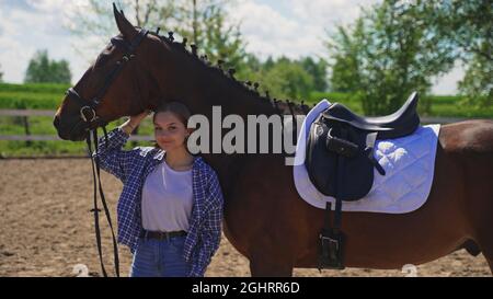 Ein junges Mädchen in einem nicht geknöpften Hemd in einem weißen T-Shirt steht unter dem Hals eines Pferdes und kratzt sich am Kopf. Hochwertige Fotos Stockfoto