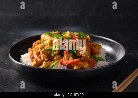 Thailändisches Gericht Gai Pad Pongali mit Huhn, Eiern, würziger, gelber thai-Paprikapaste, Tomaten, Reis in schwarzer Schüssel im Wok. Nahaufnahme. Traditionelle lokale Pause Stockfoto