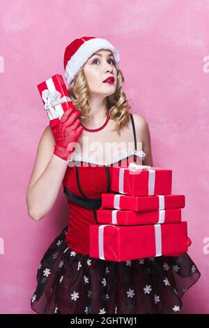 Überrascht Frau in Neujahrskleidung hält eine Menge von Boxen mit Geschenken. Rosa Hintergrund Stockfoto