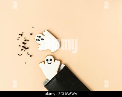 Minimale Halloween-Komposition mit Lebkuchengespenstern und schwarzen Zuckerstreuern, die schwarze Papiertüte auf beigem Hintergrund streuen. Halloween c Stockfoto