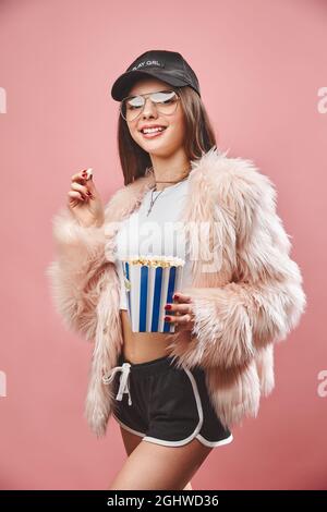 Attraktive Brünette Mädchen in faux rosa Fell hält Popcorn in der Hand. Tragen in schwarzen Shorts weißen oberen schwarzen Kappe und Brille. Blick auf die Kamera Stockfoto