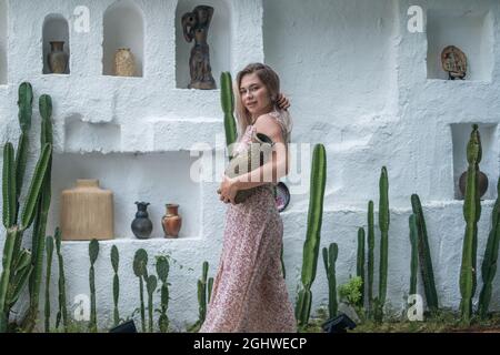 Schöne Frau mit blonden langen Haaren in langen Kleid posiert mit alten Vase in der Hand. Glückliches Gesicht. Weiße Designer-Wand mit Kakteen und Statuen Stockfoto