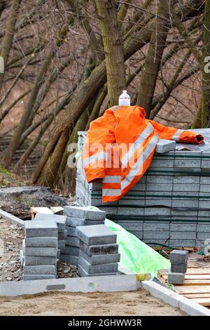 Die orangefarbene Jacke eines Baumeisters liegt auf einem Haufen Pflasterplatten vor dem Hintergrund wachsender Bäume im Park. Vertikales Bild. Stockfoto