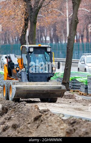 Der kompakte Straßengrader ebnet sandigen Boden vor den ankommenden Pflasterplatten entlang der Straße. Vertikales Bild. Speicherplatz kopieren. Stockfoto