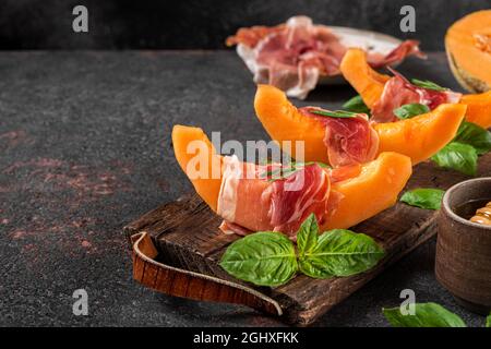 Prosciutto-Schinken mit Melone-Cantaloupe-Scheiben, Honig und Basilikum auf Schneidebrett auf dunklem Hintergrund. Italienische Vorspeise. Gesunde Ernährung Stockfoto