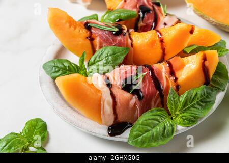 Melone-Cantaloupe in Scheiben mit Prosciutto-Schinken, Balsamico-Essigsauce und Basilikum auf einem Teller auf weißem Hintergrund. Italienische Vorspeise. Nahaufnahme. Gesunde Foo Stockfoto