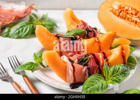 Schinken mit Melone-Cantaloupe-Scheiben, Balsamico-Essigsauce und Basilikum auf einem Teller mit Gabeln auf weißem Hintergrund. Italienische Vorspeise. Nahaufnahme. Stockfoto