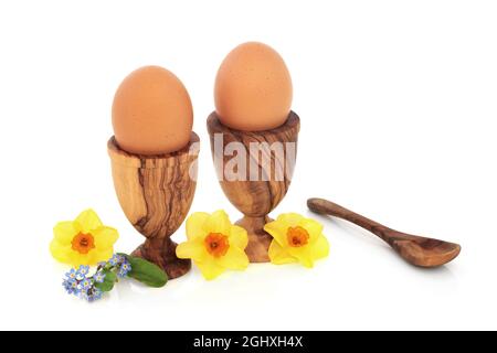 Gesunde, frische braune Eier zum Frühstück in Eierbechern aus Olivenholz mit Narzissen und Vergiss mich nicht mit Blumen. Health Food Konzept für Ostern und Frühling. Stockfoto