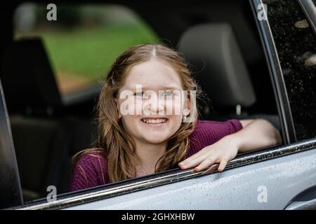 Kleines kaukasisches Mädchen mit Sommersprossen und einem lila Hemd, das aus einem Autofenster schaut Stockfoto