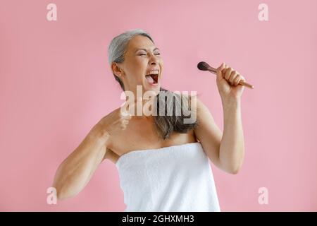 Aufgeregt mittleren Alters asiatische Modell mit Frottee Handtuch gewickelt singt auf rosa Hintergrund Stockfoto