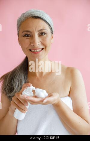 Positive asiatische Dame mittleren Alters, eingewickelt mit einem weichen Handtuch, hält Reinigungsschaum auf rosa Hintergrund Stockfoto
