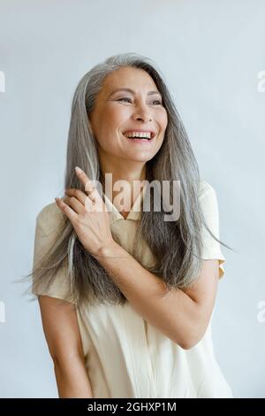 Asiatische Frau mittleren Alters berührt natürliche heidnerische Haare auf hellgrauem Hintergrund Stockfoto