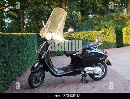 Amersfoort, Niederlande - September 3 2021. Ein schwarzer Vespa-Roller mit Windschutzscheibe, der auf einem leeren Parkplatz mit grünen Hecken abgestellt ist Stockfoto