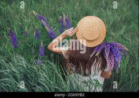 Schöne romantische Frau mit Blumenstrauß von Lupinen freudig in weißem Kleid und Hut sitzt in Feld von lila Lupine Blumen. Weicher, selektiver Fokus Stockfoto