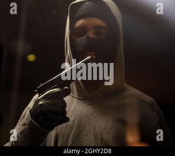 Nach dem Verbrechen Rauch aus Pistole blasen. Hier ist das Ende. Mann in schwarzer Balaclava mit grauem Kapuzenpullover, der die Waffe vor dem Mund hält. Bewaffneter Verbrecher mit pi Stockfoto