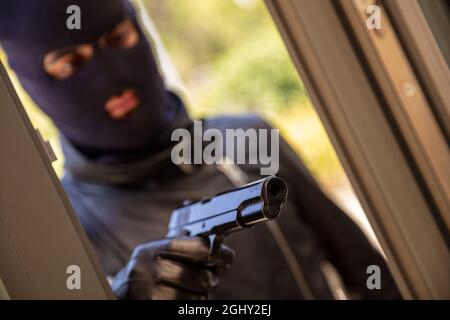 Mann in schwarzer Balaclava, der Pistole mit Lederhandschuh im Handhintergrund hält. Blur mit Kapuze bewaffneter Räuber zielt mit Waffe hinter dem Fenster. Mörder Dieb Mord Stockfoto