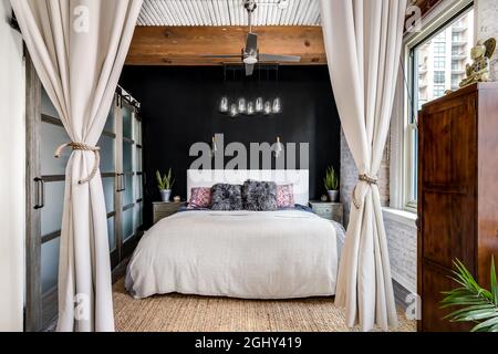 Ein Schlafzimmer in einer Loft-Wohnung mit einer freiliegenden Holzdecke, Glasschiebetüren zu einem Schrank, einer schwarzen Akzentwand und einem schicken Glaslicht, das darüber hängt. Stockfoto