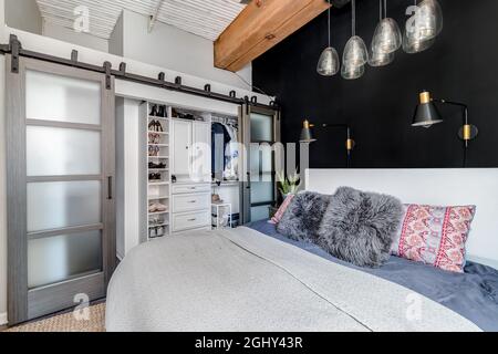 Ein Schlafzimmer in einer Loft-Wohnung mit einer freiliegenden Holzdecke, Glasschiebetüren zu einem Schrank, einer schwarzen Akzentwand und einem schicken Glaslicht, das darüber hängt. Stockfoto