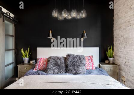 Ein Schlafzimmer-Detail, das mit einer schwarzen Unfallwand, montierten schwarzen und goldenen Lampen, einem schicken Glaslicht und farbenfrohen Kissen aufgenommen wurde. Stockfoto