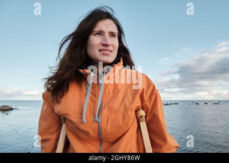 Eine Frau auf Krücken läuft am Ufer des Sees entlang. Sandstrand vor dem Hintergrund eines ruhigen Sees und Wolken am Horizont Stockfoto