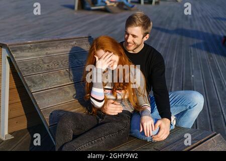 Ein junges verliebter Mann, ein Mann und eine Frau umarmen sich auf einer Bank im Erholungsgebiet. Ein schönes rothaariges Mädchen in Pullover und Jeans, ein attraktiver junger Mann mit blonden Haaren. Das glückliche Paar. Stockfoto