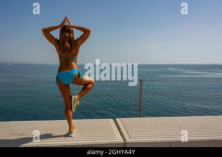 Erwachsene Frau, die einen zweiteiligen Bikini trägt und Yoga auf einem Dock am Meer macht Stockfoto