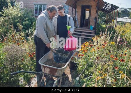 Ein 30-jähriger Mann gießt Wasser in eine Zementmischung, um Zement für das Ausgießen von Wegen in einem Garten zu schaffen Stockfoto