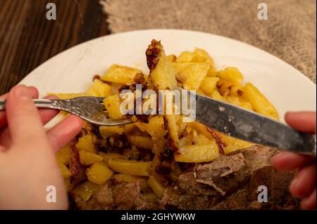 Jemand schneidet mit einem Messer und einer Gabel ein Stück saftiges Braten auf einem Teller gebratener Kartoffeln. Nahaufnahme Selektiver Fokus Stockfoto