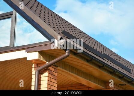 Giebel-Dachkonstruktion mit braunem Metall-Ziegeldach, Regenrinne, Schneeräumen und Dachfenster gegen blauen Himmel. Stockfoto