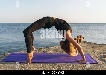 Eine Frau in schwarzer Sportkleidung, die Yoga praktiziert, führt Urdhva Dhanurasana-Übung durch, Brückenpose am Meer, stehend auf einer Matte Stockfoto