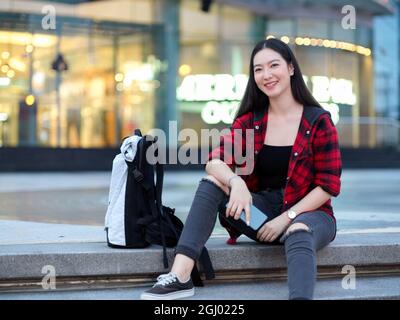 Attraktive asiatische Hipster junge Frau Alleinreisende mit Rucksack sitzt auf der Treppe vor dem Stadtzentrum Einkaufszentrum in der Nacht, Mädchen posiert Stockfoto