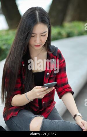 Teenager, die auf der Straße sitzt, Nachrichten an ihre Freunde gibt, ihr Smartphone nutzt, um Zeit zu verschwenden, in den sozialen Medien scrollt Stockfoto