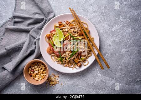 Draufsicht auf Teller mit asiatischen udon-Nudeln, würzigen Garnelen, gebratenem Tofu-Käse, Erdnüssen, Limette und grünen Zwiebeln auf grauem Betongrund Stockfoto