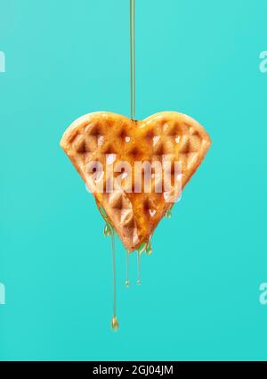 Gießen von Ahornsirup über eine herzförmige Waffel vor einem grünen Hintergrund. Nahaufnahme von Ahornsirup, der auf eine Waffel tropft. Stockfoto