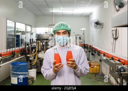 Junger asiatischer Arbeiter in steriler Uniform, der das Produkt aus abgefülltem rotem Saft in der Produktionslinie in der Getränkeverarbeitungsfabrik zeigt Stockfoto