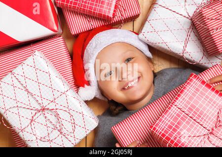 Draufsicht auf ein kleines Mädchen, das einen weihnachtsmütze trägt. Das Kind liegt auf dem Boden, umgeben von Kisten mit Weihnachtsgeschenken Stockfoto