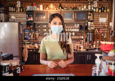 Attraktive junge asiatische Frau Unternehmer trägt Gesichtsmaske stehen Kunden an Holztheke im Restaurant begrüßen Stockfoto