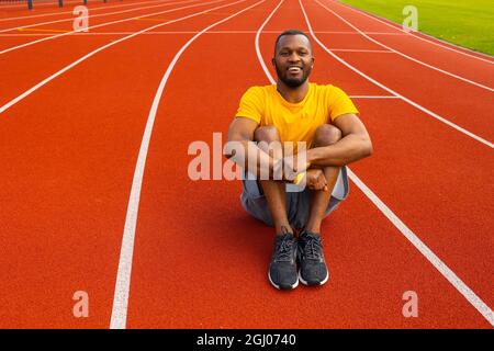 Porträt eines afroamerikanischen Athleten, der nach dem Laufen auf der Stadionstrecke sitzt. Sportlicher Mann im gelben T-Shirt nimmt eine Pause vom Training ein Stockfoto