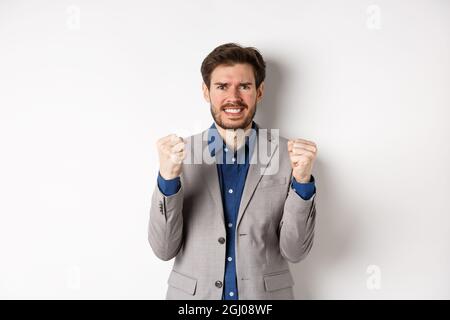Angespannter Geschäftsmann, nervös und unter Druck, mit Fäusten und Zähnen, starrend auf die Kamera, Geld verdienen und besorgt aussehen, auf Weiß stehend Stockfoto