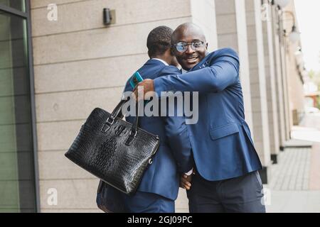 Porträt von zwei schwarzen afroamerikanischen Geschäftsleuten in Anzügen, die sich die Hände schüttelten und sich im Freien eng umarmten. Die Freude, gute Freunde zu treffen Stockfoto
