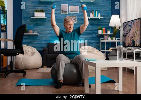 Fokussierter Rentner auf Fitness sitzen schweizer Ball heben Hand streching Arm Muskel tun Gesundheitsübungen. Ältere Frau, die im Wohnzimmer mit Hanteln Körperwiderstand trainiert Stockfoto
