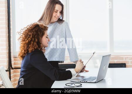 Schöne junge Brünette Frau und rothaarige Frau reden und diskutieren Arbeitsmomente mit Laptop im Büro. Stockfoto