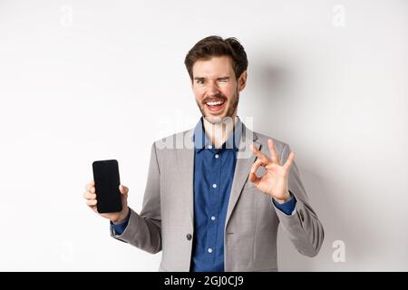 E-Commerce und Online-Shopping-Konzept. Erfolgreicher Geschäftsmann Geld verdienen auf dem Smartphone, zeigt leeren Telefonbildschirm und okay Zeichen, zwinkert glücklich an Stockfoto