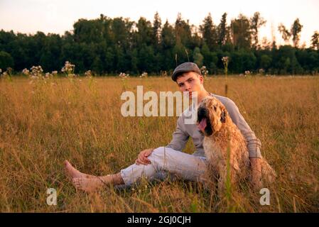 Ein 18-jähriger Junge mit Mütze sitzt an einem Sommerabend mit einem Hund auf einem Feld im dicken Gras. Stockfoto