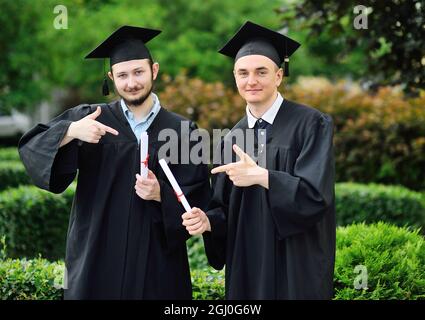 Zwei junge Hochschulabsolventen in Gewändern und quadratischen Hüten freuen sich über ein Diplom. Stockfoto