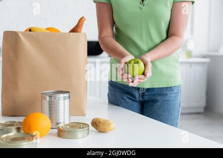 Beschnittene Ansicht einer Frau, die einen Apfel in der Nähe von Essen und Papiertüte in der Küche hält Stockfoto