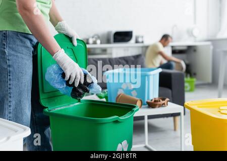 Beschnittene Ansicht einer Frau in Latexhandschuhen, die Plastiktüten mit Recycling-Schild in den Papierkorb legt Stockfoto