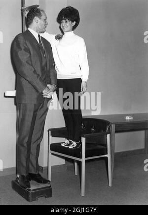 Club Instructioness Monique Farhi, die 2 cm groß ist, klettert auf einem Stuhl, um die Höhe von Henry Cooper, dem britischen Schwergewicht-Boxchampion, der 2 cm groß ist, zu überprüfen. 8. März 1966 Stockfoto