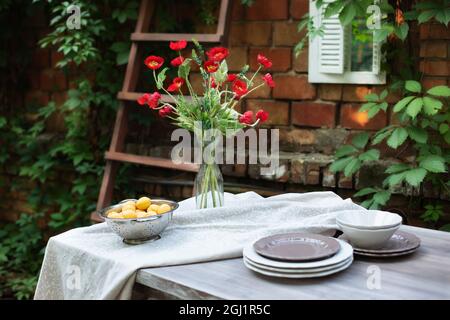 Ein Blumenstrauß aus rotem Mohn in einer Glasvase. Blumen in einer Vase zu Hause. Innenterrasse, Terrasse. Holztisch und Teller für Gartenparty oder Abendessen Stockfoto